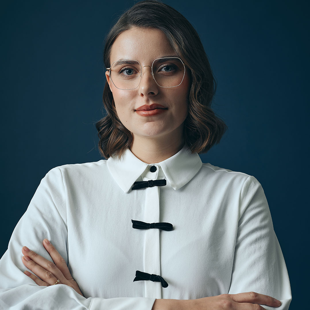 Frau mit heller Brille und weißer Bluse vor dunkelblauem Hintergrund.