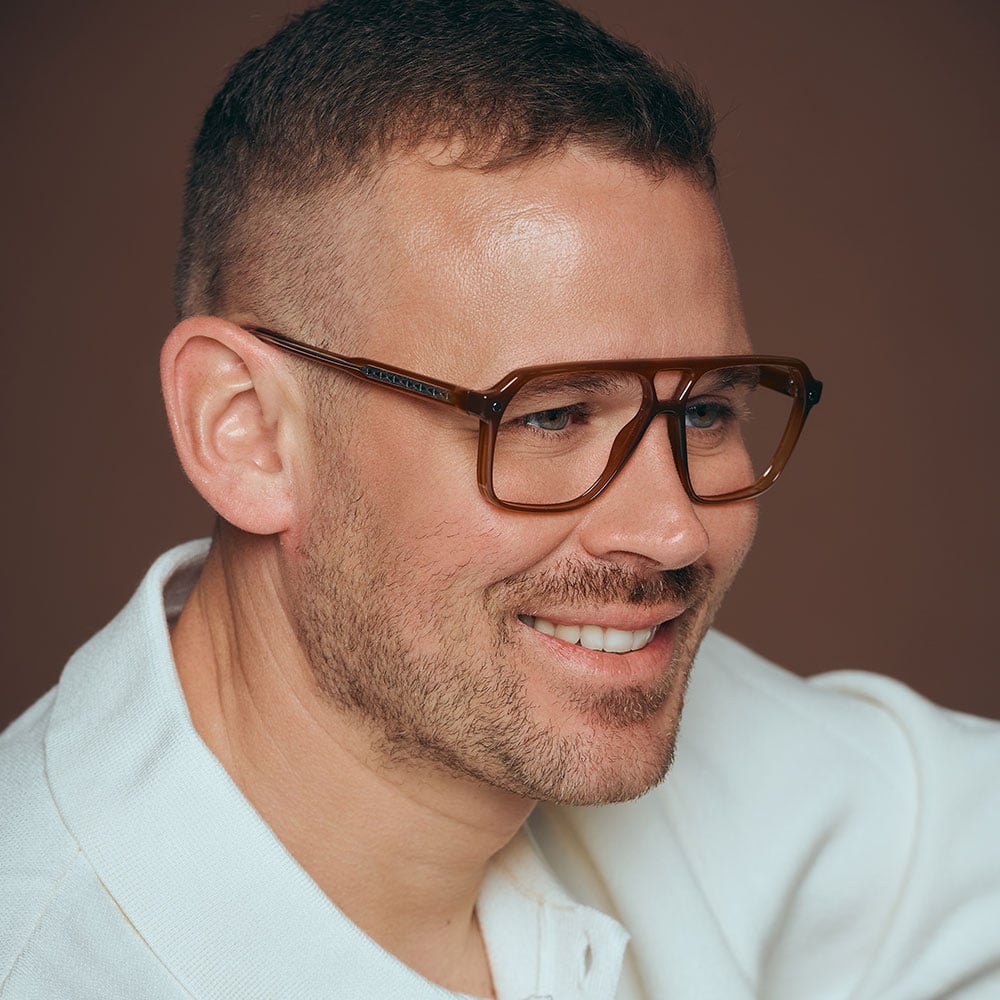 Lächelnder Mann mit brauner Brille und hellem Hemd vor braunem Hintergrund.